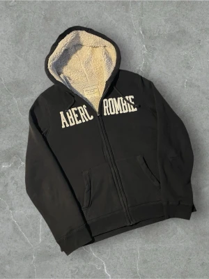 Svart hoodie från Abercrombie med päls | L - Svart hoodie med dragkedja från Abercrombie. Stor vit logga över bröstet och två fickor framtill. Insidan av huvan är fodrad med mjuk beige teddy, vilket gör den extra mysig. Klassisk passform och ribbade muddar vid ärmar och nederkant.