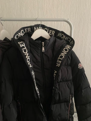 Svart dunjacka från Moncler - Snygg svart dunjacka från Moncler med stor huva och tydlig logga längs kanten. Jackan har dragkedja framtill, två fickor med dragkedja och klassisk Moncler-patch på ärmen. Puffig modell som håller värmen och ger en trendig look. 15-16 years. 34-36 storlek.