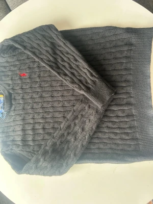 Mörkgrå kabelstickad tröja Polo Ralph Lauren - Mörkgrå kabelstickad tröja från Polo Ralph Lauren med klassisk röd logga broderad på bröstet. Tröjan har rund halsringning och ribbade muddar vid ärmslut och nederkant. Perfekt för dig som gillar stilren och tidlös design.