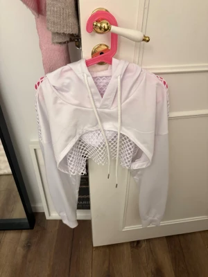 Vit cropped mesh hoodie - Säljer en vit cropped hoodie med huva och dragsko. Tröjan har långa ärmar och coolt mesh-tyg framtill och nedtill, vilket ger en sportig och trendig vibe. Perfekt för dig som gillar streetwear och vill sticka ut lite extra.
