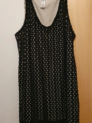 Klänning - Svart virkad klänning från H&M med ärmlös design och rund hals. Klänningen är kort och har ett vitt knytband i midjan som detalj.  Materialet är syntet och modellen har ett öppet, nätliknande mönster.