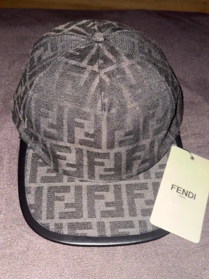 Grå/svart Fendi keps med FF-mönster - Snygg grå keps från Fendi med det klassiska FF-logomönstret över hela kepsen. Skärmen är svart och har en lätt böjd form. Kepsen är tillverkad i tygmaterial och har en modern streetwear-vibe. Perfekt för dig som vill sticka ut med en exklusiv accessoar.