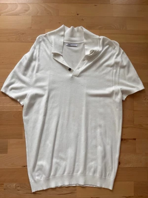 Vit polotopp från Zara med knappar - Säljer en stilren vit polotopp från Zara med korta ärmar och två knappar vid halsen. Toppen har en mjuk och lätt stickad känsla och passar perfekt för dig som gillar en clean look. Snygg krage och ribbad nederkant ger en modern touch.