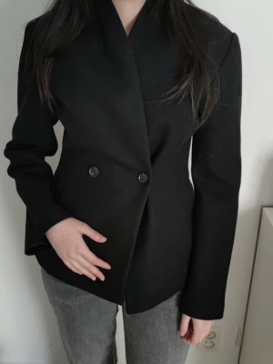 Svart kavaj med dubbelknäppning - Stilren svart kavaj med dubbelknäppning framtill och klassisk krage. Kavajen har en figurnära passform och långa ärmar. Ny med prislapp!
