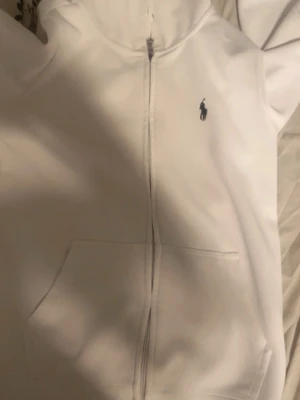 Vit hoodie från Polo Ralph Lauren - Klassisk vit hoodie från Polo Ralph Lauren med dragkedja framtill och fickor på magen. Märkets ikoniska logga i mörkblått broderad på bröstet. Tillverkad i mjukt material och har huva för extra komfort.