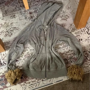 Grå hoodie med fuskpälsdetaljer - Unik grå hoodie med dragkedja och stor huva. Ärmsluten har coola detaljer i brun fuskpäls som ger en lekfull och trendig look. Hoodien har fickor framtill och ribbade muddar. Perfekt för dig som vill sticka ut lite extra.