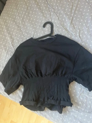 Svart croppad topp med ribbad midja - Snygg svart croppad topp med rund hals och korta ärmar. Toppen har en ribbad, åtsittande midja som ger en cool siluett och markerar midjan. Perfekt till höga jeans eller kjol för en trendig look.