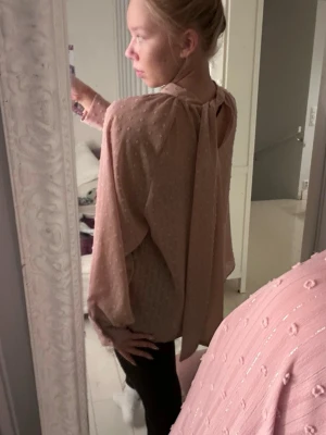 Rosa blus med prickig struktur - Säljer en ljusrosa blus med långärmad design och dekorativa små prickar i tyget. Har en knytdetalj, går att ha på en rosett eller bara hängande band i ryggen. 