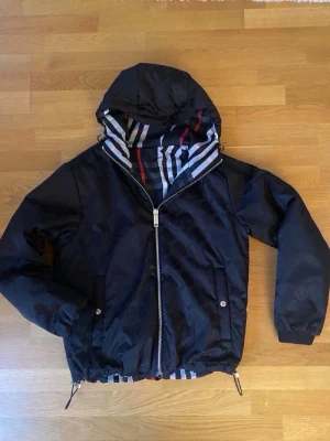 Svart vindjacka från Burberry - Snygg svart vindjacka från Burberry med ikoniskt rutmönster i vitt, rött sop grått på insidan och baksidan. Jackan har huva, dragkedja framtill, två fickor med tryckknappar och justerbar nederkant. Materialet är lätt och vindtätt, perfekt för blåsiga dagar.