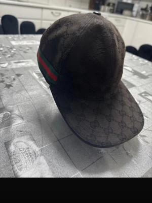 Gucci keps med GG-mönster - Säljer en svart keps från Gucci med klassiskt GG-monogram över hela kepsen och den ikoniska gröna och röda randen på sidan. Kepsen har böjd skärm och är tillverkad i ett slitstarkt tygmaterial. Perfekt för dig som vill ha en stilren och lyxig accessoar.