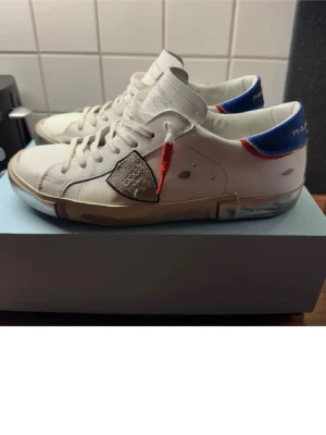 Philippe Model vita sneakers med blå detalj - Snygga HELT NYA vita sneakers från Philippe Model med blå och röda detaljer på hälen och en grå patch på sidan. Skorna har klassisk låg profil, vita snören och transparent sula. Perfekta för dig som vill ha en stilren och unik look.