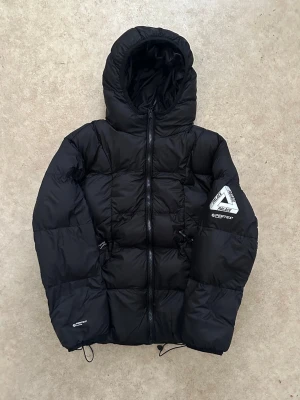 Svart dunjacka från Palace - Svart dunjacka från Palace med stor huva och dragkedja framtill. Jackan har Palace-logga på ärmen och på huvan, samt justerbar dragsko nedtill. Puffig modell i klassisk pufferstil, perfekt för kalla dagar. Kommer med tag!