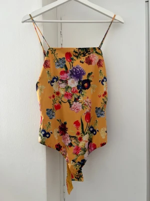 Gul blommig body med smala axelband - Snygg gul body med färgglatt blommönster i rött, lila, blått och grönt. Modellen har smala axelband och hög skärning vid benen. Materialet ser ut att vara mjukt och följsamt, perfekt för en trendig look. Inget synligt märke.