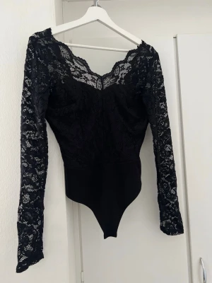 Svart spetsbody med långa ärmar - Snygg svart body med långa ärmar i genombruten spets och v-ringad hals. Överdelen är helt i spets med blommönster och nederdelen i slät stretchig textil. Perfekt för dig som vill ha en elegant och trendig look. Knäppning i grenen för enkel på- och avtagning.