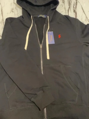 Ralph lauren hoodie  - Snygg svart hoodie från Polo Ralph Lauren med dragkedja, vit dragsko och klassisk röd logga på bröstet. Tillverkad i mjuk bomull och har två fickor framtill samt ribbade muddar vid ärmslut och nederkant. Perfekt för en chill och stilren look.
