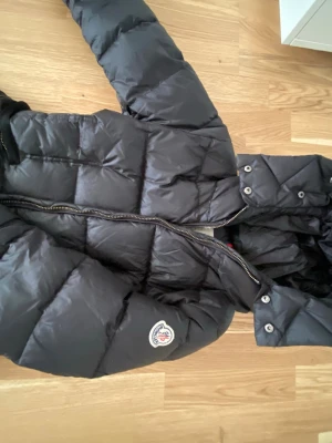 Svart dunjacka från Moncler - Säljer en svart dunjacka från Moncler med klassisk quiltad design och huva. Jackan har dragkedja framtill, tryckknappar vid kragen och Moncler-logga på ärmen. Insidan har den ikoniska serieteckningen. Perfekt för kalla vinterdagar.
