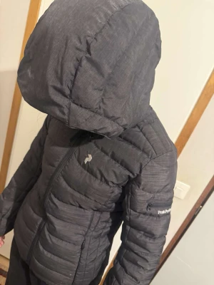 Svart dunjacka från Peak Performance - Snygg svart dunjacka från Peak Performance med huva och dragkedja framtill. Jackan har en quiltad design, diskret logga på bröstet och ärmarna samt figurnära passform. Perfekt för kalla dagar och enkel att matcha med olika outfits.