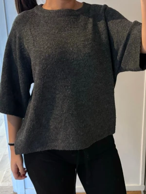Mörkgrå stickad tröja med halvärm - Mysig mörkgrå stickad tröja med avslappnad passform och halvlånga ärmar. Tröjan har rund halsringning och är tillverkad i ett mjukt, tjockare material som passar perfekt till kyligare dagar. Enkel och stilren design som är lätt att matcha.