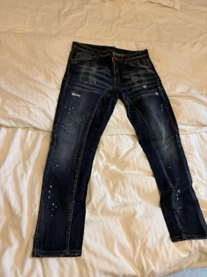 Mörkblå distressed jeans från Dsquared2 - Säljer ett par mörkblå jeans från Dsquared2 med coola slitningar, färgstänk och en relaxed fit. Jeansen har klassiska fem fickor, orange patch bak och kontrastsömmar. Perfekta för dig som gillar en streetwear-look.(A Kopia)