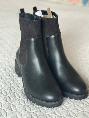 Svarta Chelsea boots med grov sula - Stilrena svarta Chelsea boots med elastiska ribbade partier på sidorna och chunky gummisula. Boots i slätt konstläder med rund tå och låg klack. Perfekta för dig som vill ha en klassisk men trendig look. Från bonprix. Storlek 43 men jag tycker dom verkar mer som en 42. Endast provat på hemma men har legat och blivit lite dammiga och det finns liten repa på ena. Se bild. Nypris 499. Köpt utan box 