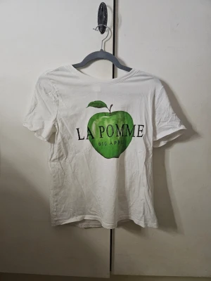 Vit t-shirt med grön äppleprint - Säljer en jätte söt vit t-shirt från H&M i storlek S med stort grönt äpple och texten 'LA POMME BIG APPLE' på framsidan. Klassisk rund hals och korta ärmar. Tillverkad i mjuk bomull. Är använd men har inga tecken på fläckar, skador eller andra slitage. Säljer den då den tyvärr inte längre kommer till användning. Nypris runt 150 kr. 💓