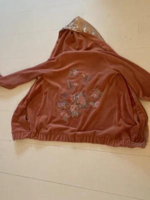 Rostbrun hoodie med broderi - Säljer en mjuk hoodie i rostbrunt sammetstyg med broderade blommor och fåglar på ryggen. Hoodien har huva och resår i nederkanten. Perfekt för dig som gillar unika detaljer och skön stil.