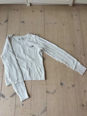 Vit kabelstickad tröja Hollister - Kabelstickad vit tröja från Hollister med V-ringning och lång ärm. Tröjan har ribbade muddar och nederkant samt en liten broderad logga i vinrött på bröstet. Perfekt för en avslappnad men snygg look.