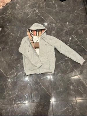 Grå zip hoodie från Burberry - Snygg grå zip hoodie från Burberry med klassiskt rutigt foder i huvan. Tröjan har dragkedja framtill, två fickor och ribbade muddar vid ärmslut och nederkant. Perfekt för en avslappnad och stilren look.