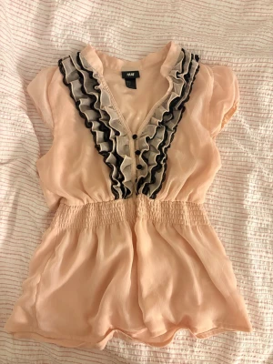 Ljusrosa volangblus från H&M - Söt ljusrosa pepblumtopp/blus från H&M med svarta och beige volangdetaljer längs knäppningen framtill. I strl S. 🌷🌷
