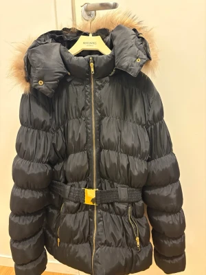 Svart pufferjacka med fuskpäls - Snygg svart jacka, tung jacka , med guldfärgade dragkedjor, avtagbar huva med fuskpäls och bälte i midjan. Jackan har quiltad design och ger en riktigt cozy vinterlook. Perfekt för kalla dagar när du vill vara både varm och stilren.