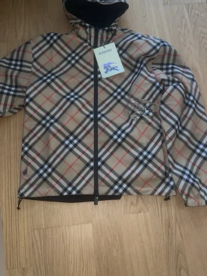 Burberry beige rutig vindjacka - Snygg beige vindjacka från Burberry med klassiskt rutmönster i svart, vitt och rött. Jackan har huva, hel dragkedja framtill och en diskret Burberry-logga på bröstet. Tillverkad i ett lätt och vindtätt material, perfekt för blåsiga dagar.