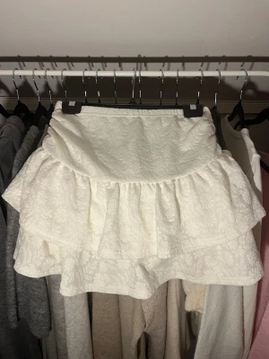 Vit volangkjol med blommönster - Supersöt vit kjol med volanger och blommönster. Kjolen har inbyggda shorts och en stretchig midja.