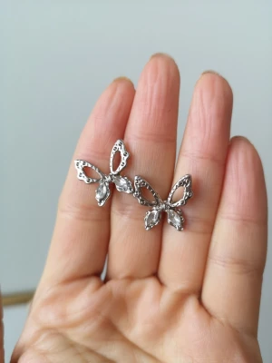 Silvriga fjärilsörhängen med stenar - Snygga örhängen i form av fjärilar i silverfärg med gnistrande klara stenar på vingarna. Örhängena har en öppen design och detaljerade kanter som ger en elegant look. Perfekta för dig som gillar unika accessoarer med lite extra bling.