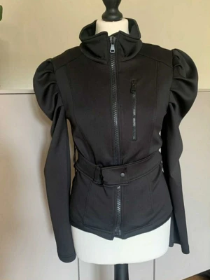 Svart jacka med puffärmar och bälte - Stilren svart jacka med markerade puffärmar, hög krage och bälte i midjan. Jackan har dragkedja framtill och en extra ficka med dragkedja på bröstet. Snygg, figurnära passform och coola detaljer som ger en unik look.