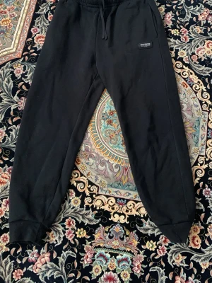 Svarta mjukisbyxor från McKenzie - Säljer ett par svarta mjukisbyxor från McKenzie med resår i midjan och dragsko. Byxorna har ribbade muddar vid bensluten och en diskret logga på vänster sida. Perfekta för chill dagar och streetwear-stil.