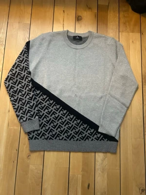 Grå stickad fendi tröja - Snygg grå stickad tröja från Fendi med diagonal svart detalj och ikoniskt FF-mönster på ena ärmen och nedre delen. Rund halsringning och ribbade muddar. Perfekt för dig som gillar exklusiv streetwear och vill sticka ut.