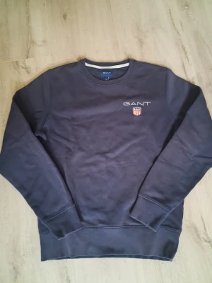 Mörkblå sweatshirt från GANT - Säljer en mörkblå sweatshirt från GANT i storlek S. Tröjan har rund hals, ribbade muddar och ett broderat GANT-logo med sköld på bröstet. Materialet är mjuk bomull och passformen är normal. Perfekt för chill dagar och snygg till jeans.