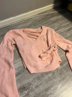 Ljusrosa kofta från lager 157 - Supersöt ljusrosa stickad kofta från 157 med knytband i sidan och v-ringad hals. Tröjan har långa ärmar och ribbade muddar vid ärmslut och nederkant. Perfekt för dig som gillar en feminin och trendig look.