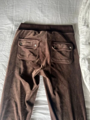 Bruna Juicy Couture byxor med fickor - Mysiga bruna mjukisbyxor i mjukt velourmaterial med bred resår i midjan och dragsko. Byxorna har två stora fickor fram och två bakfickor med lock och knappdetaljer. Perfekta för chill dagar hemma eller när du vill vara bekväm.