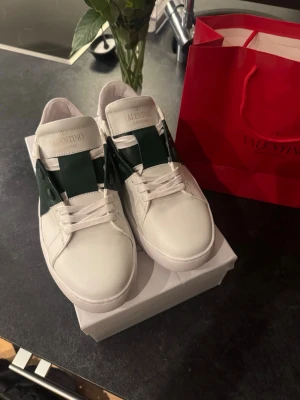 Valentino vita gröna sneakers - Snygga vita sneakers från Valentino med en bred grön läderdetalj över snörningen och klassisk logga på plösen. Skorna har en låg profil och subtila nitar på hälen för extra edge. Perfekta för dig som vill ha en clean men unik look.  Har inte haft domma länge men under ett par månader kvitto kommer med 