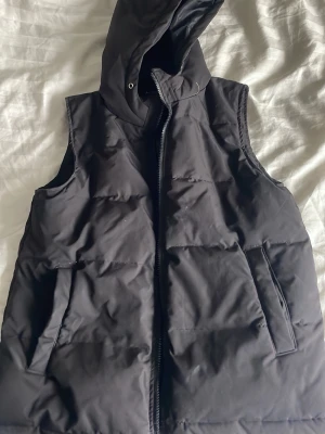 Svart dunväst med huva - Säljer en svart dunväst med huva och dragkedja framtill. Västen har två stora fickor på sidorna och en stilren, quiltad design. Perfekt för lager-på-lager och ger en cool look till din outfit. Går inte att stänga 