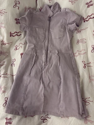 Ljuslila jeansklänning med dragkedja - Kort jeansklänning i ljuslila med markerad krage, två bröstfickor och dragkedja framtill. Klänningen har korta ärmar, barnstorlek 152.