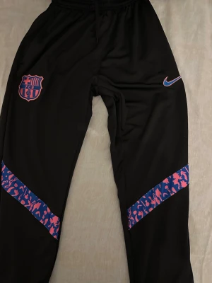 Barcelona Nike träningsbyxor svart - Snygga svarta träningsbyxor från Nike med FC Barcelona-logga på vänster ben och Nike Swoosh på höger. Byxorna har färgglada mönstrade detaljer i blått och rosa vid knäna. Perfekta för fotboll eller gymmet.