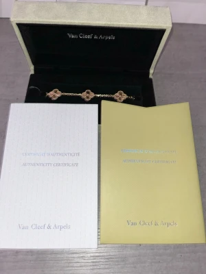 Van Cleef & Arpels Alhambra armband - Exklusivt Alhambra armband från Van Cleef & Arpels med tre fyrklöverformade länkar i rosa sten och guldfärgad metall. Armbandet har en tunn kedja och levereras i originalask med äkthetsintyg. Perfekt för dig som gillar lyxiga och tidlösa smycken.