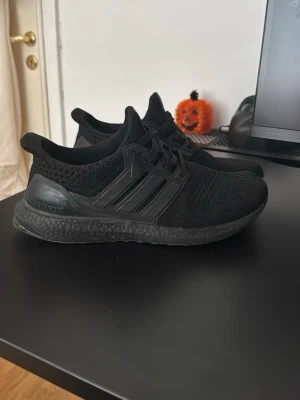 Svarta Adidas Ultraboost sneakers - Säljer ett par helsvarta Adidas Ultraboost sneakers med stickad ovandel och Boost-sula för maxad komfort. Skorna har klassiska tre ränder på sidan, snörning och en sportig look som passar perfekt till streetwear. Materialet är mesh och syntet.