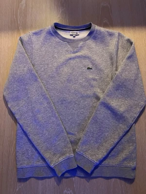 Grå sweatshirt från Lacoste - Klassisk grå sweatshirt från Lacoste med rund halsringning och diskret krokodillogga på bröstet. Tröjan har långa ärmar, ribbade muddar och är tillverkad i mjukt bomullsmaterial. Perfekt för en avslappnad och stilren look.