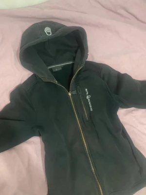 Svart hoodie från Sail Racing - Svart hoodie från Sail Racing med dragkedja framtill och en extra ficka med dragkedja på bröstet. Märkets logga syns både på bröstet och på huvan. Hoodien har en klassisk passform och är tillverkad i mjukt material.