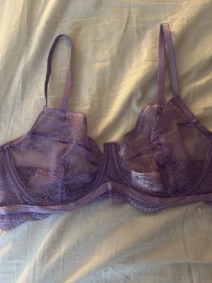 Lila spets-bh med tunna band - Säljer en lila bh i transparent spets med tunna axelband och dekorativ kant. Bh:n har bygel och vacker blommig spetsdetalj över hela kupan. Perfekt för dig som gillar en feminin och trendig look.