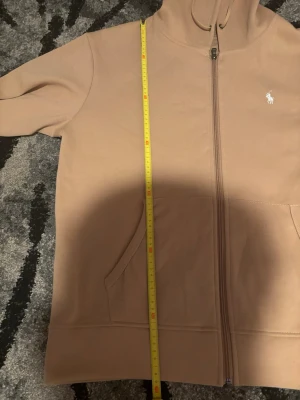 Beige hoodie från Polo Ralph Lauren - Snygg beige hoodie från Polo Ralph Lauren med dragkedja framtill och klassisk vit logga på bröstet. Tröjan har huva med dragsko och två fickor framtill. Perfekt för en avslappnad och stilren look.