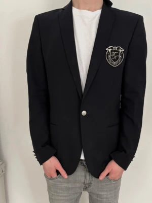 Svart kavaj med emblem från SikSilk - Stilren svart kavaj från SikSilk med silverfärgat emblem på bröstet och knappdetaljer vid ärmslut. Klassisk passform med smala slag och enkelknäppning. Perfekt för en dressad look. Från Flippa K